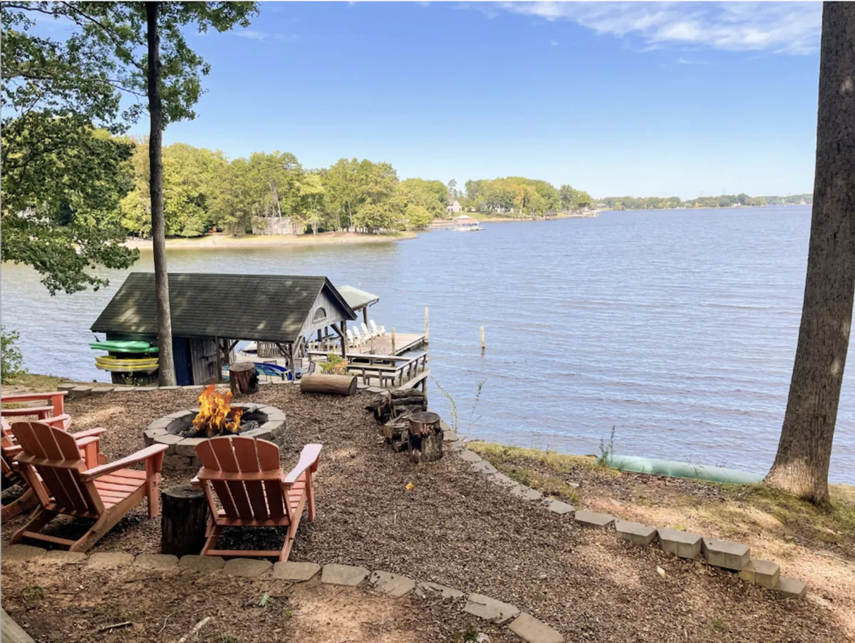 This exterior image showcases a welcoming fire pit surrounded by Adirondack chairs, with an inviting lake view and a boathouse in the background. The setting suggests the perfect spot for campfire gatherings, making s'mores, and enjoying sunsets over the water.