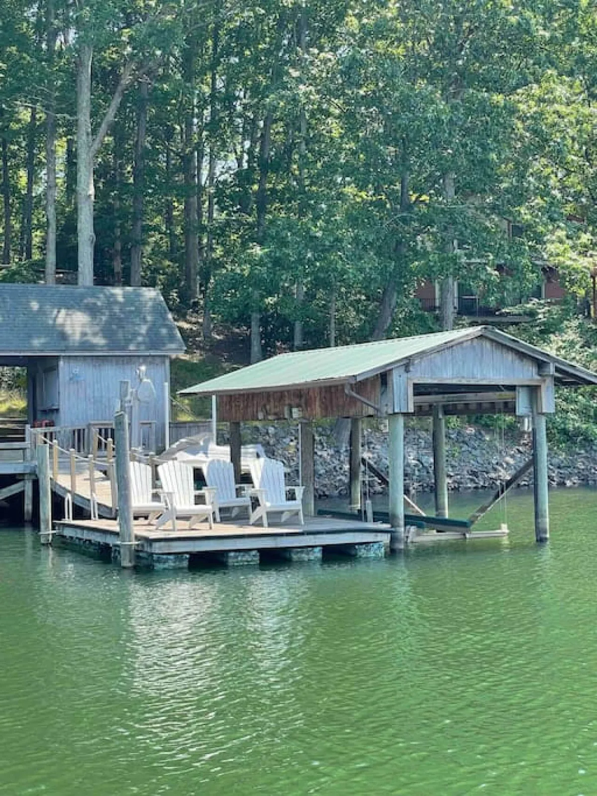 A dock extends over the water with adirondack chairs positioned for relaxation, and a boathouse nearby. The lake is glassy, reflecting the early morning or dusk light, and the setup is perfect for small gatherings or solo reflection before or after water adventures. The image captures the essence of lakeside living and leisure.