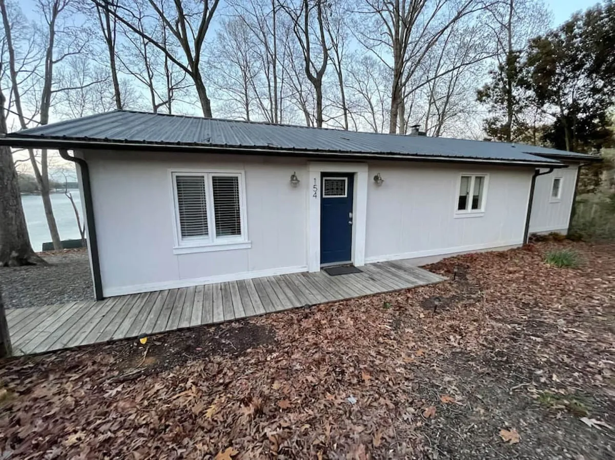 The front entrance of the property features a striking blue door, large windows, and is surrounded by mature trees. The setting feels calm and inviting, with the deck area offering views of the yard and natural surroundings—an ideal spot for guests and their pets to relax after a day of lakeside adventure.