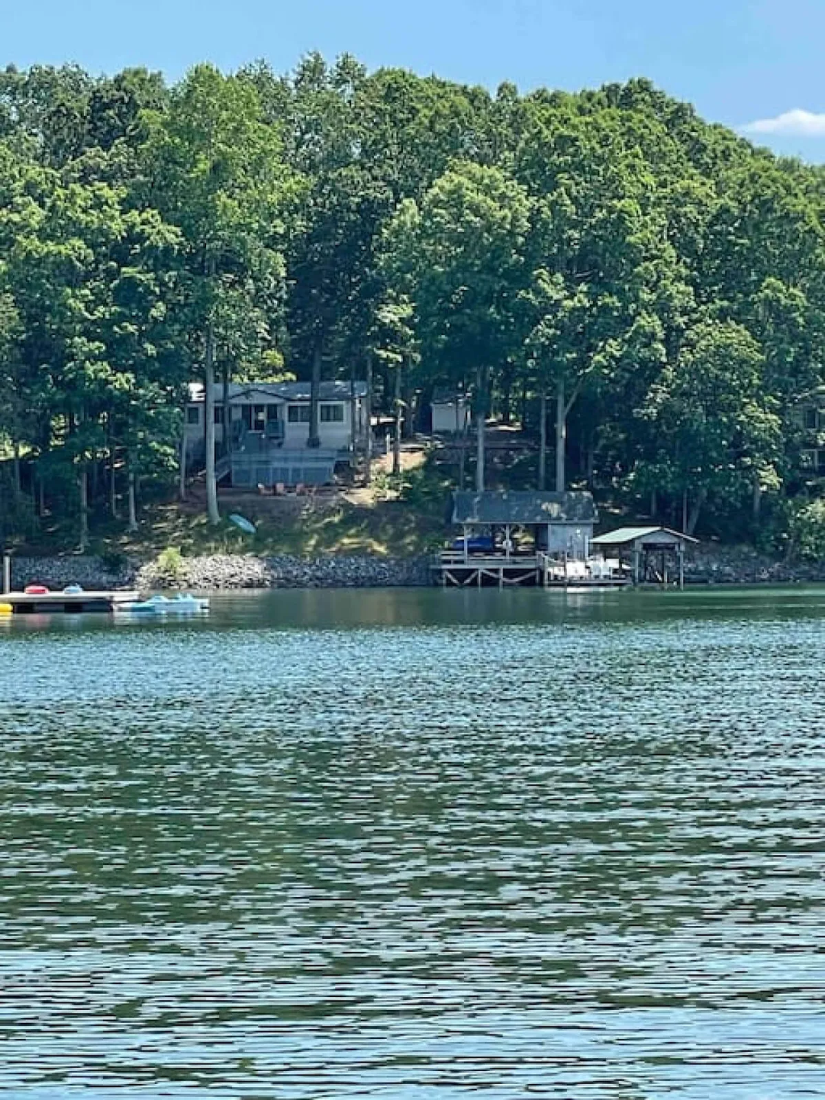 A tranquil lake stretches out in the foreground, bordered by a lush treeline. A charming house with a welcoming deck sits nestled among the trees, while a dock extends into the calm water, inviting quiet mornings and serene afternoons by the lake.