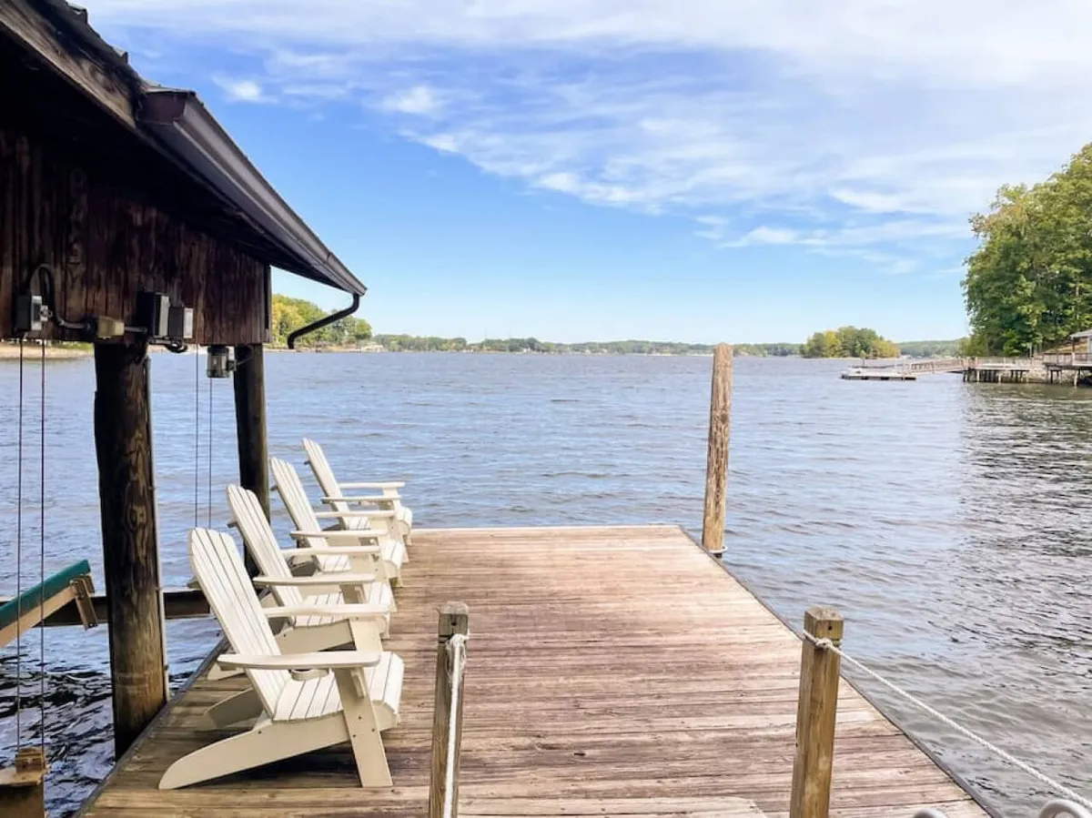 A classic wooden dock extends over calm lake waters, with Adirondack chairs positioned for relaxing and soaking in the sunrise or sunset. The dock is surrounded by lush trees, creating a peaceful, private setting—perfect for launching kayaks or paddle boards, sunbathing, or enjoying a quiet drink by the water.