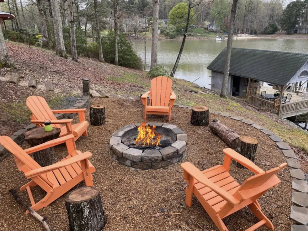 A cozy outdoor fire pit surrounded by bright orange Adirondack chairs, set among trees with a beautiful view of the lake in the background. The setup is ideal for campfire gatherings, storytelling, or simply enjoying a peaceful evening by the water. The warm ambiance invites groups to gather and connect.