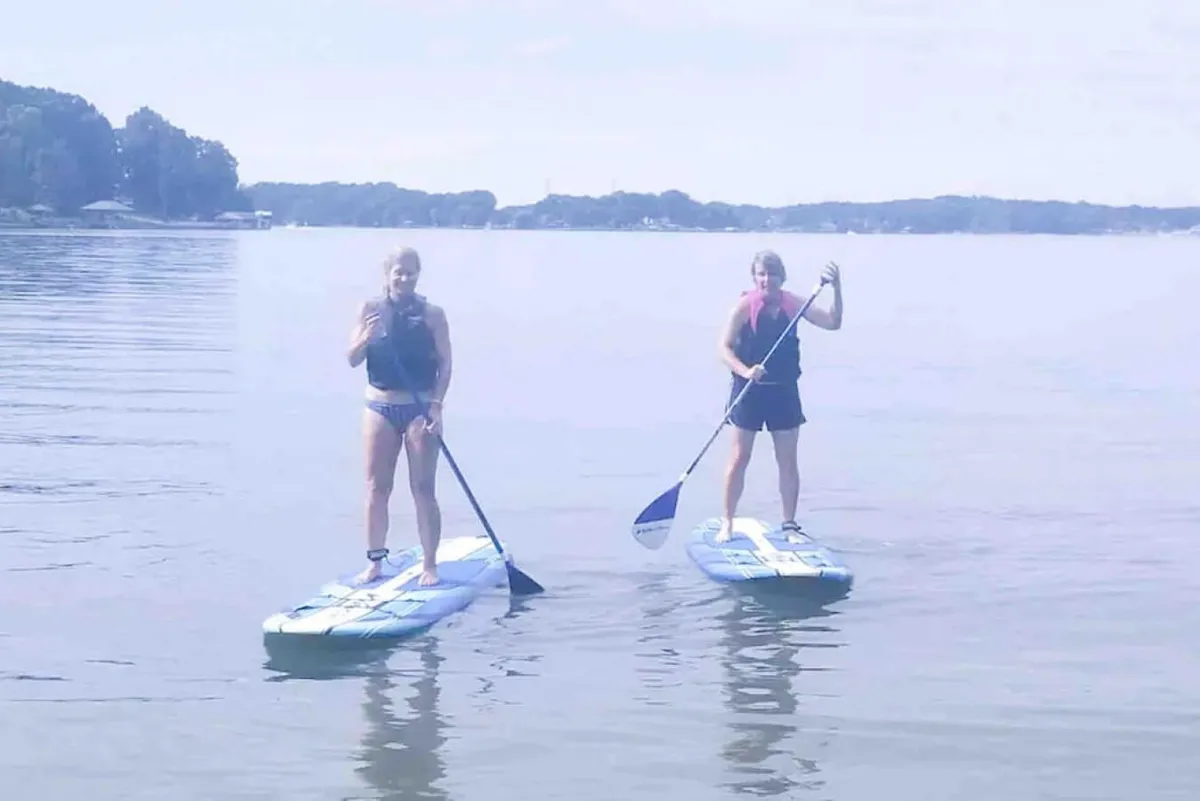 Two women paddleboarding on the calm lake, with a distant shoreline and clear skies overhead. The image exudes a sense of adventure, exploration, and joy, showing the fun and active side of lakeside living and the accessibility of water sports from the property.