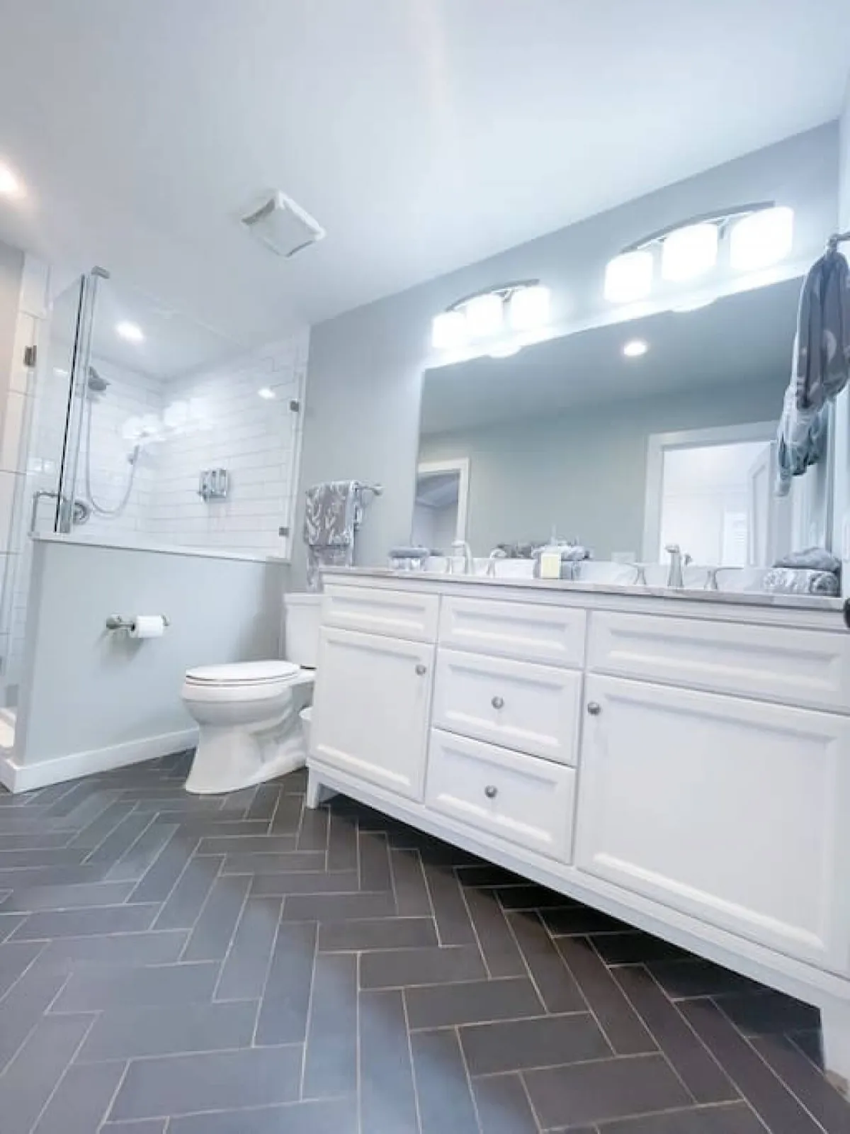 A serene bathroom featuring a double-sink vanity, a glass-enclosed shower, and calming, neutral décor. The herringbone tile flooring adds a touch of sophistication, and the room exudes a soothing, spa-like vibe.