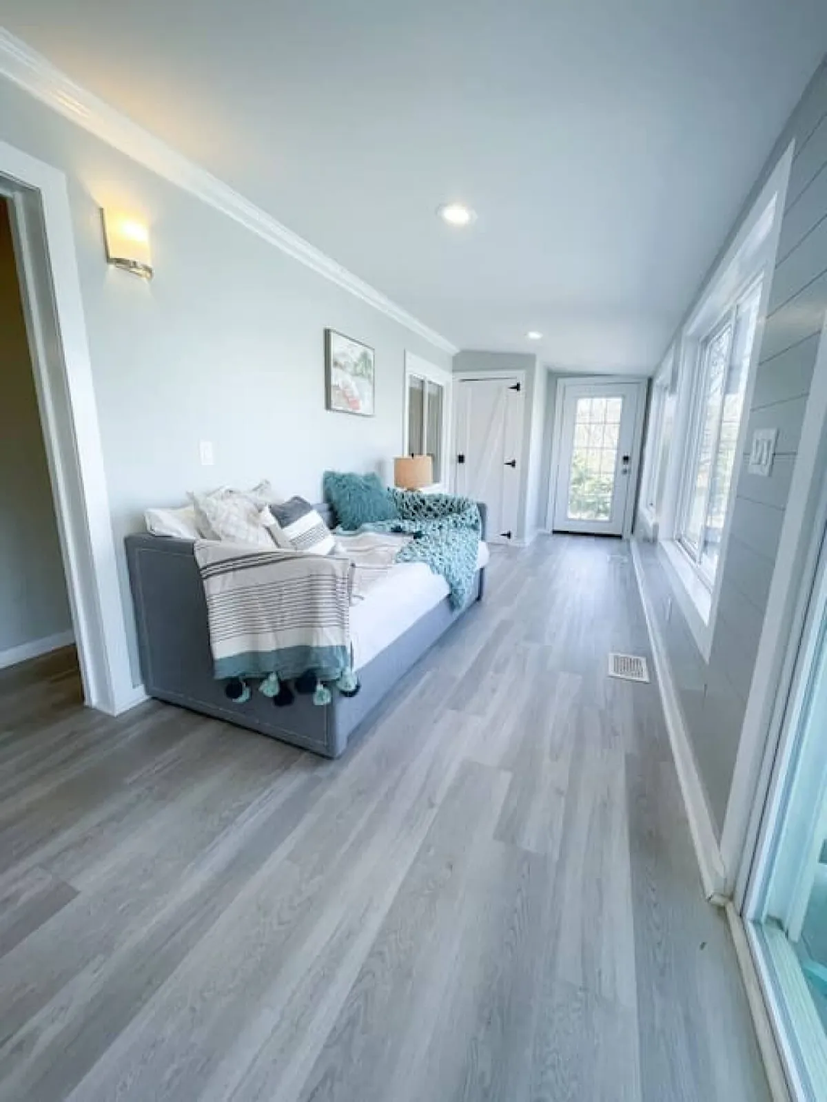 A cozy living area with a plush sofa adorned with throw pillows, a lamp casting warm light, framed artwork, and large windows that flood the room with natural light. The space feels both serene and inviting, perfect for relaxing or socializing.