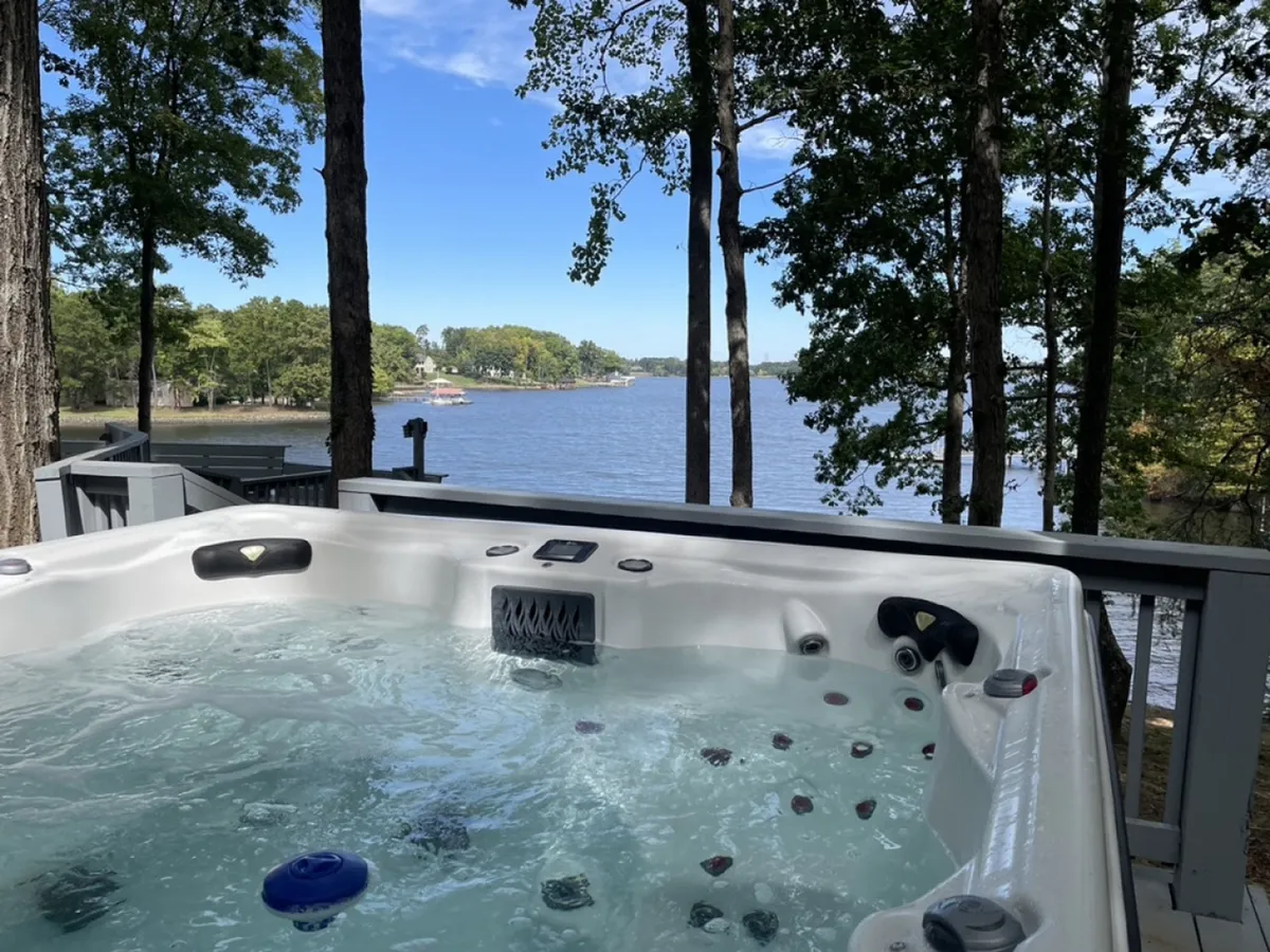 The image displays a tranquil lakeside setting with a bubbling hot tub overlooking expansive views of Lake Norman, framed by lush trees. The hot tub is positioned to maximize sunset vistas, inviting relaxation and conversation. The natural scenery is calm and inviting, suggesting privacy and a connection to nature.
