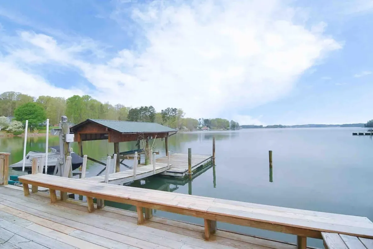 A sweeping lake view from the dock, framed by tall trees and open sky. Boating activity is visible in the distance, and the dock is ready for launching watercraft or simply soaking in panoramic views. The mood is serene, inviting both adventure and relaxation.