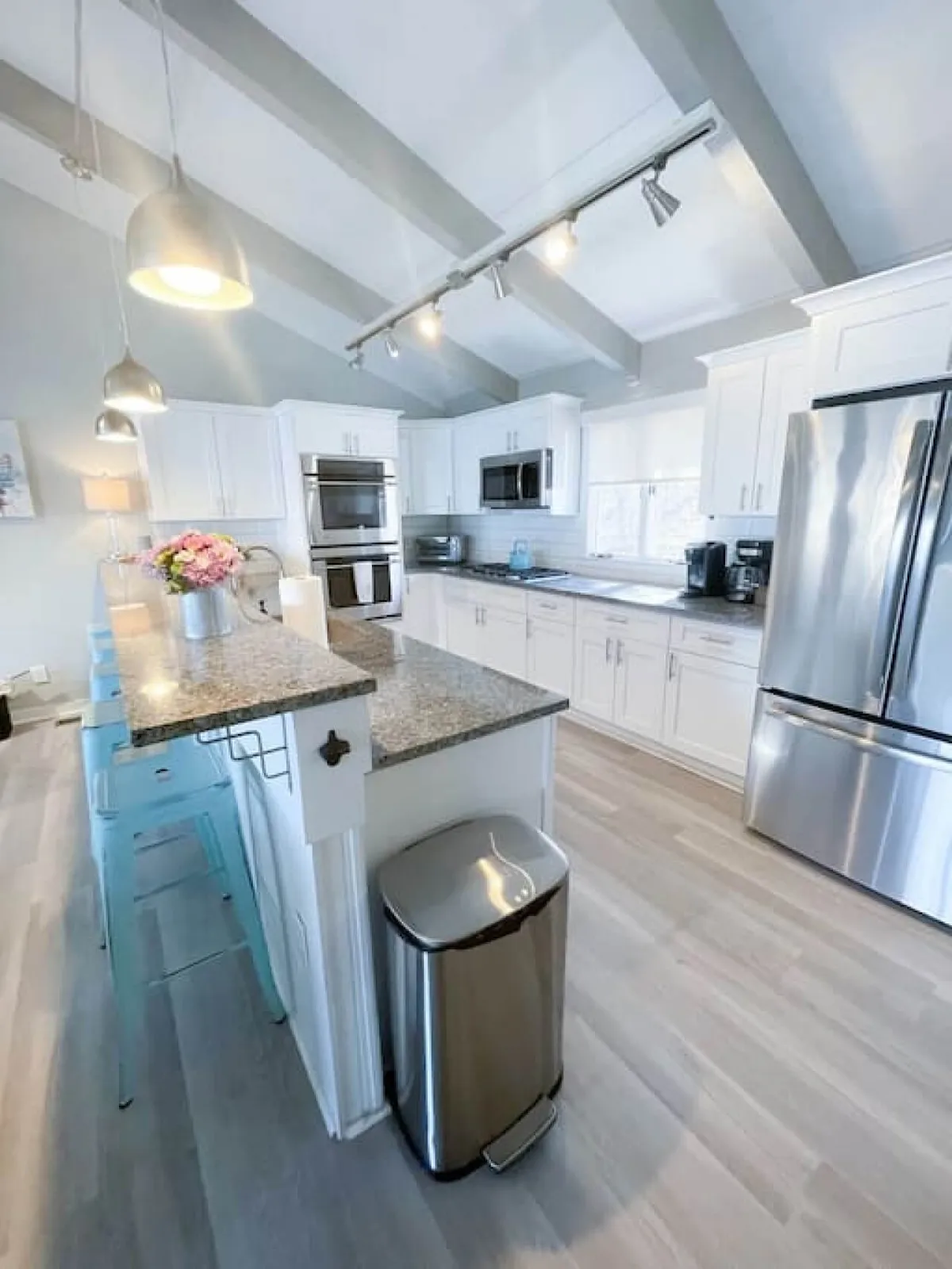 A spacious, modern kitchen featuring granite countertops, stainless steel appliances, elegant light fixtures, and blue bar stools around a central island. A vase of fresh flowers adds a welcoming touch, and the overall layout is designed for both entertaining and everyday living.