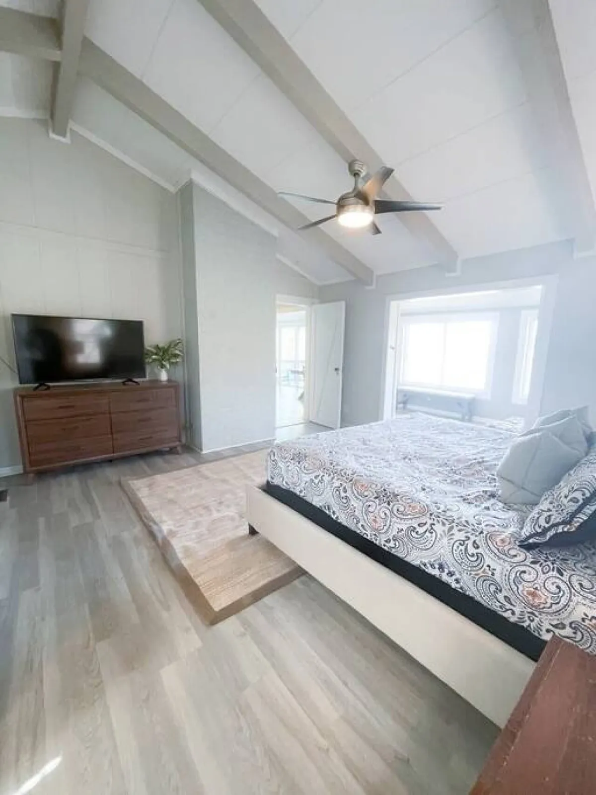 A cozy bedroom showcasing a bed with patterned bedding, a wooden dresser, a ceiling fan, and a large window that allows natural light to flood the room. The scene feels intimate and relaxing, perfect for unwinding.
