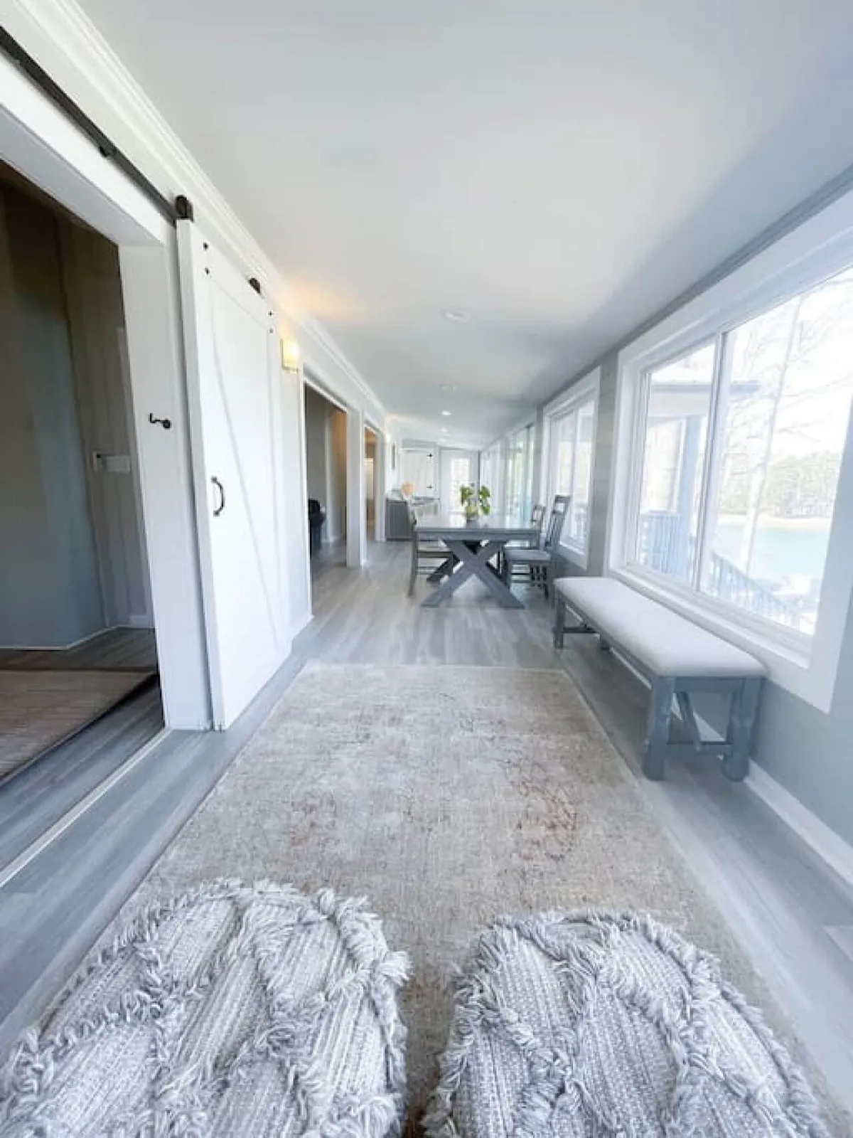 A long hallway leads to an inviting dining area with a large table and comfortable seating, set beside expansive windows that let in plenty of sunlight. The scene suggests a place for family meals, casual gatherings, and relaxed, homey moments.