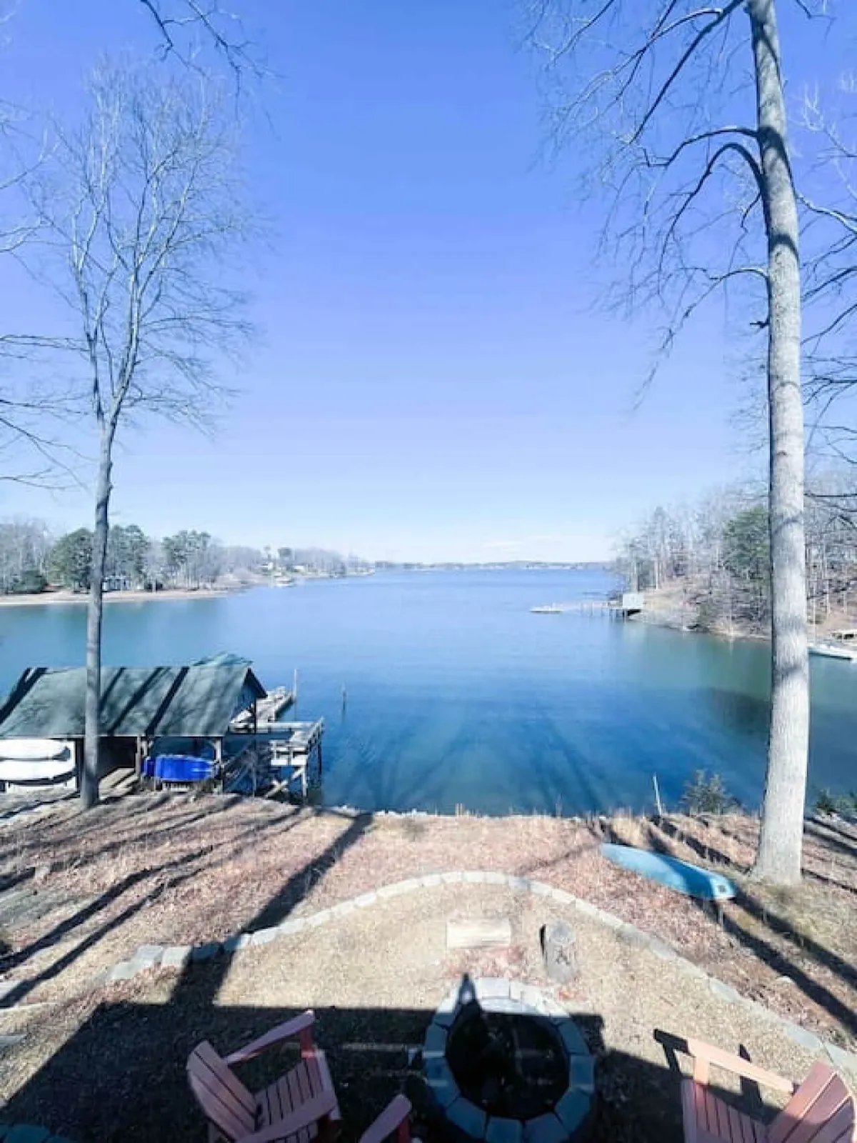 This image captures the exterior of the property with a tranquil lake, bare trees, a fire pit, and a dock with a boathouse. The spacious yard leads directly to the water, providing plenty of room for dogs to play and explore. The serene atmosphere is highlighted by sunset reflections and early morning mist.
