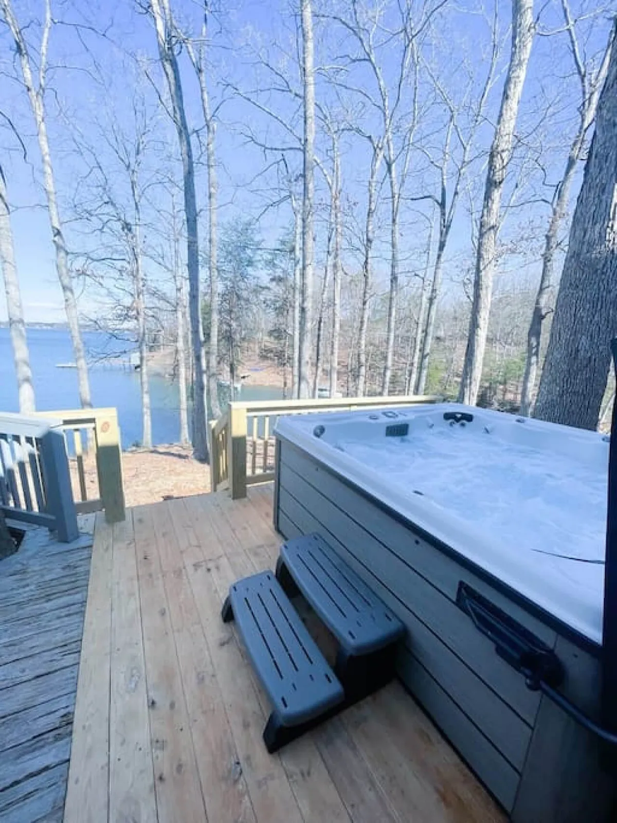 A serene scene of a wooden deck overlooking the lake, featuring a steaming hot tub nestled under bare trees. The deck offers unobstructed views of the water, inviting guests to relax and unwind in the warmth of the hot tub while enjoying the tranquil surroundings. The setting is ideal for socializing, stargazing, or simply soaking in the peaceful atmosphere.